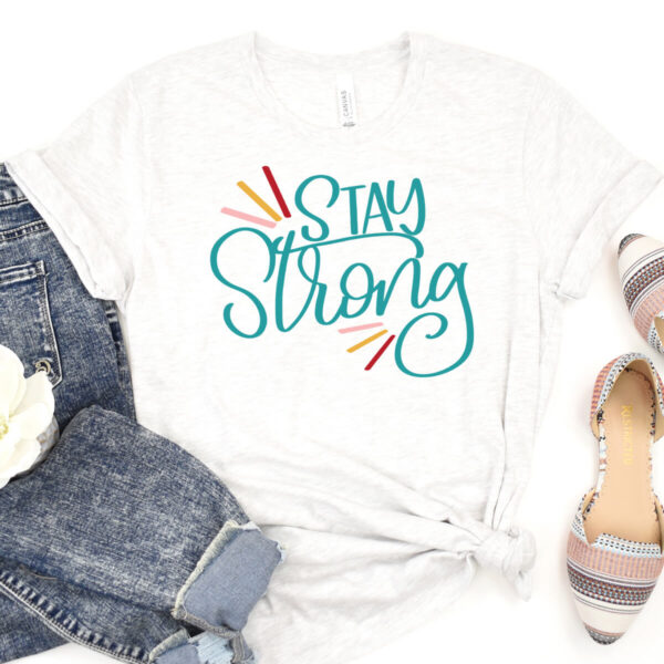 Jeans, slip-on shoes, and a white t-shirt that says "Stay Strong" lay on a white backdrop. This design is available as a free SVG cut file. 