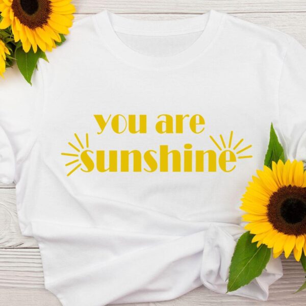 Bright yellow text on a white t-shirt says, "You are sunshine." The t-shirt is adorned with sunflowers, and the SVG cut file for the t-shirt can be downloaded for free.