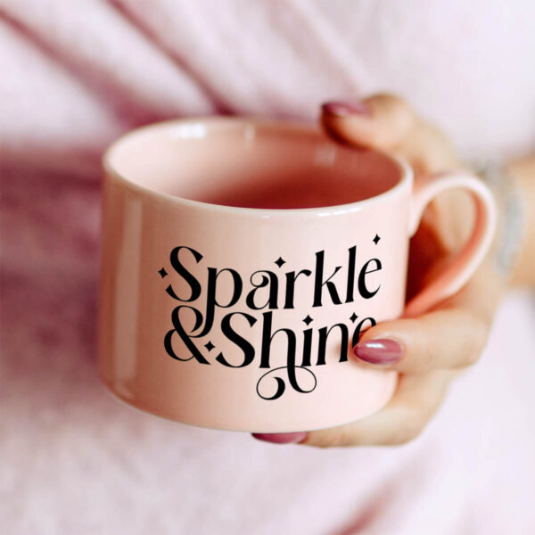 A woman's hand holds a short, wide pink mug that has been crafted with a free SVG file that says "Sparkle & Shine." 