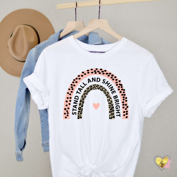 Hanging on a wooden rod next to a brown hat and in front of a denim jacket is a white t-shirt with "Stand tall and shine bright" text in a leopard and rainbow craft design.