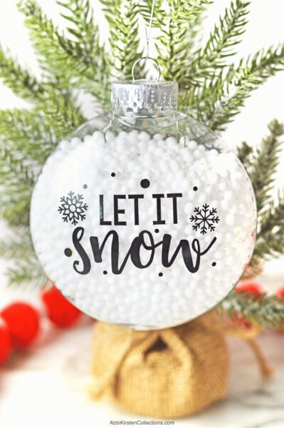 Tiny white balls rest in a clear round fillable Christmas ornament that hangs from a bough. The bauble reads "Let it Snow" with decorative vinyl snowflakes. 