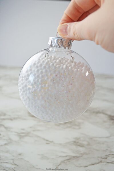 A close-up of Abbi Kirsten's hand holding the top of a fillable Christmas ornament full of white balls and glitter snow. 