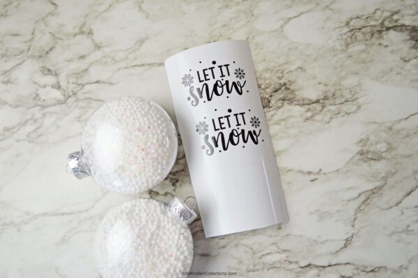 Two fillable Christmas ornaments full of small white balls lay next to a piece of vinyl that says “Let it Snow.” You can make these ornaments with this free SVG file and a Cricut cutting machine. 