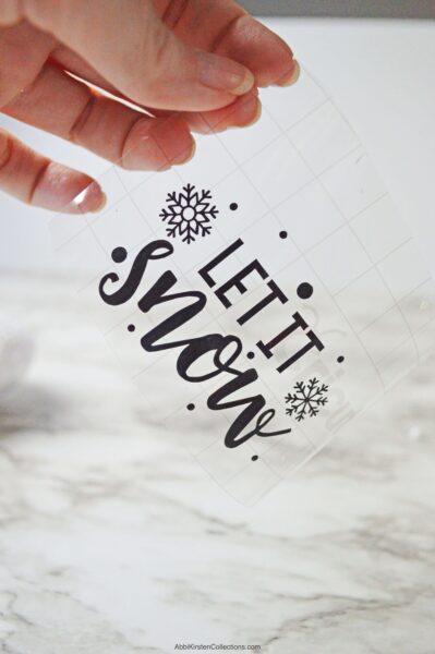 A close-up of Abbi Kirsten’s hand holding clear transfer tape with black vinyl adhered to it. The vinyl says “Let it Snow” and is a free SVG file. 