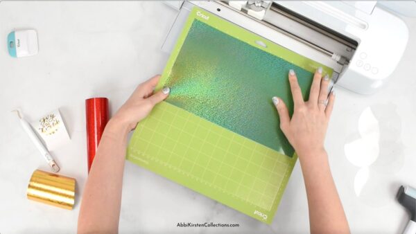 The image shows hands loading a Cricut machine with holographic green vinyl. 