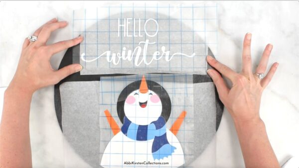 Abbi Kirsten's hands show how to align the vinyl "hello winter" snowman design on a DIY wood sign. 