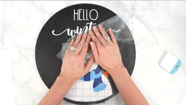 Abbi shows how to carefully remove transfer tape from the "hello winter" layered vinyl design onto the wood sign. 