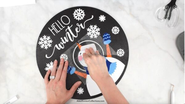 An overhead view of Abbi Kirsten's hands burnishing the final vinyl snowflakes onto the DIY circular seasonal wood sign craft. 