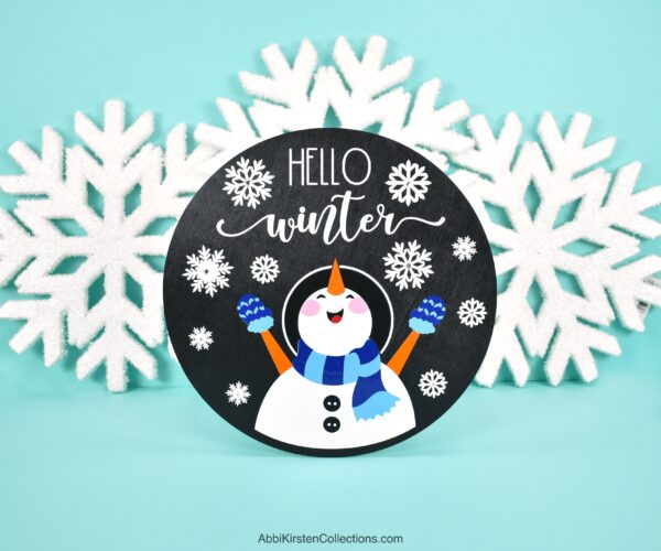 Image shows a black wood sign in front of decorative snowflakes. The sign has a snowman and the phrase "hello winter." The tutorial teaches how to layer vinyl on wood signs with your Cricut machine. 