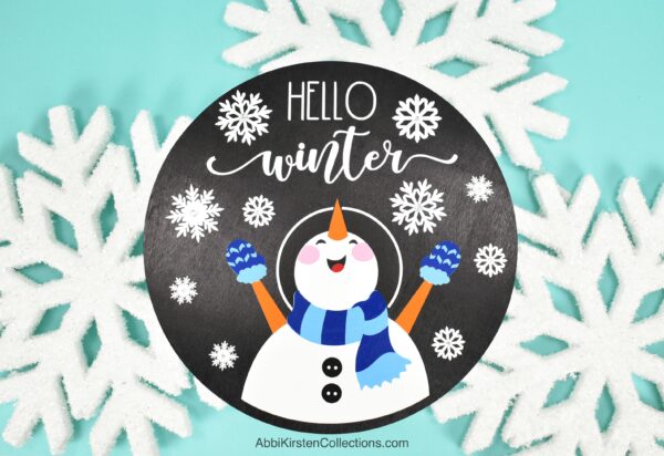 A black wood sign with a snowman and the phrase "hello winter" surrounded by big snowflake decorations. The tutorial teachers how to layer vinyl on wood signs with your Cricut machine. Tips and tricks for aligning layers of vinyl.