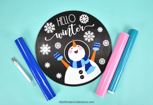 A handmade circular wood sign showing a snowman happy in snowflakes with the words "hello winter" above it. Markers and a weeding tool lay off to the side. 