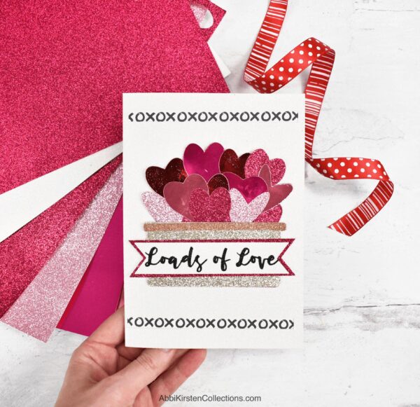 Abbi Kirsten's hand holding a Valentine's Day card that reads "Loads of Love." Red, pink and white glitter cardstock is behind the card with a decorative red ribbon. 