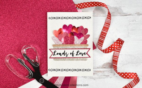 A finished, handmade Valentine's Day card with cutout glitter hearts and the text "Loads of Love" on the front, leans against a red glitter piece of cardstock adorned with red polka dot ribbon and a pair of craft scissors. 