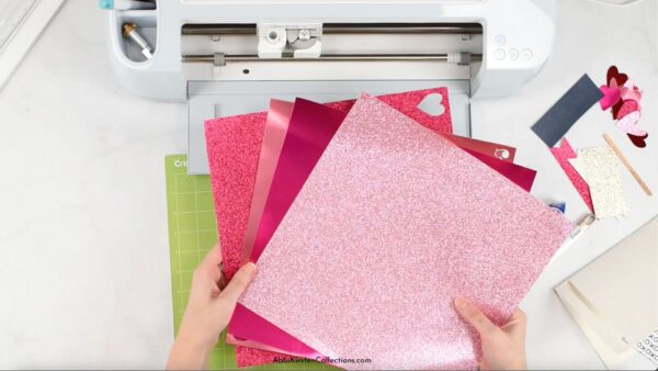 Abbi Kirsten's hands hold glitter cardstock in a fan above a Cricut cutting machine and mat. A Valentine's Day card lays to the right. 