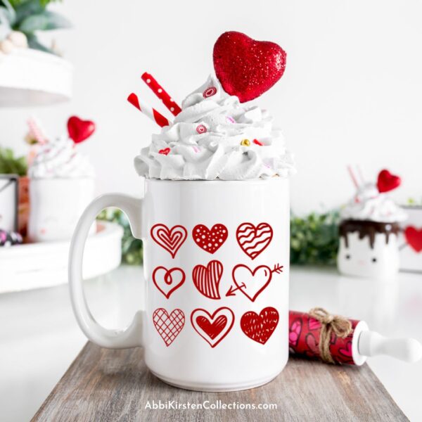 A tall white mug full of whipped cream with sprinkles, hearts and red stiped paper straws has six red hearts printed on it and was made using SVG cut files and a Cricut machine. 