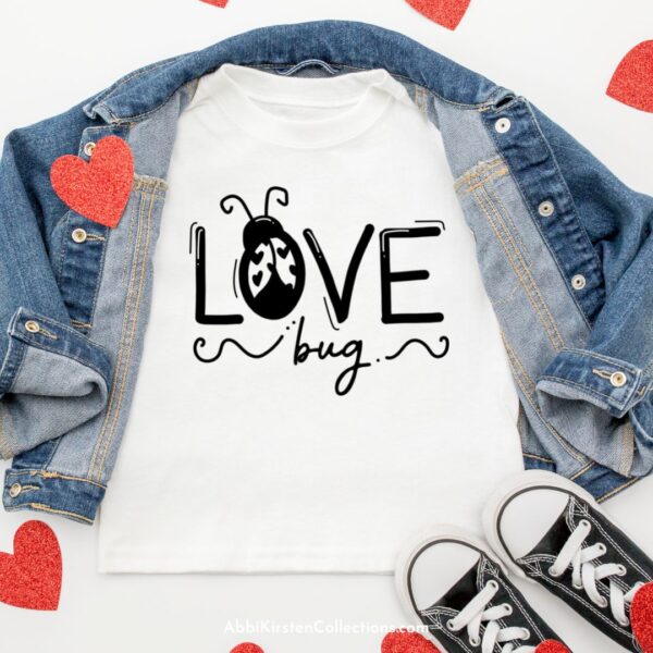 A white t-shirt folded on a white background with a jean jacket and black and white sneakers. The t-shirt has the words "Love Bug" with a ladybug as the "o." Made using a SVG file. 