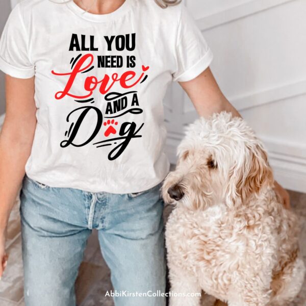 A blonde woman is photographed from her neck to her knees as she kneels next to her dog. She is wearing a white t-shirt made with a SVG cut file that says "all you need is love and a dog."