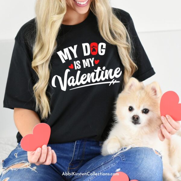 A blonde woman in blue jeans and a black T-shirt that reads "my dog is my valentine" smiles at her cute small dog while holding red paper hearts in her hands. This t-shirt was made using SVG cut files and a Cricut machine. 
