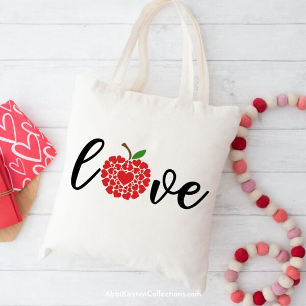 A tan tote bag with the word "love" with an apple made of hearts lays next to red, white and pink beads on a string and a part of a red and white heart package. A perfect gift for teachers on Valentine's day