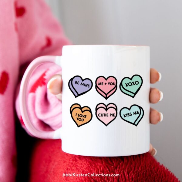 A woman's hand holds a white mug with candy hearts made with a SVG craft file.
