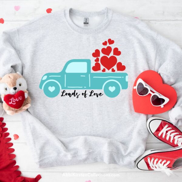 A child's sweatshirt has a blue pickup truck with hearts in the truck printed on it. A heart with sunglasses, a plush Valentine's toy, and red sneakers adorn the shirt. 
