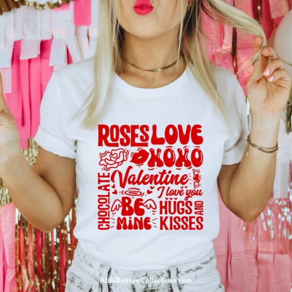 A woman with red lipstick stands in front of Valentine's Day streamers while wearing a white t-shirt printed with red Valentine's Day words, made via sublimation and a Cricut machine.