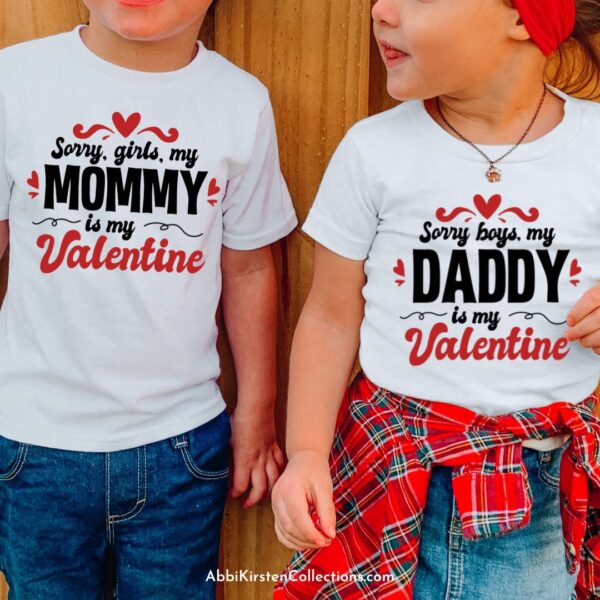 A close-up of two toddlers wearing white t-shirts against a wood wall. The t-shirts are made using a free SVG file and the boy's t-shirt reads "sorry, girls, my mommy is my Valentine," and the girl's t-shirt days "sorry, boys, my daddy is my Valentine." 