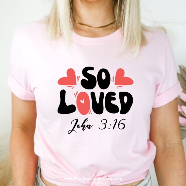 A close-up on a blonde model wearing a pink t-shirt with the bible verse "so loved John 3:16" crafted onto the front. 