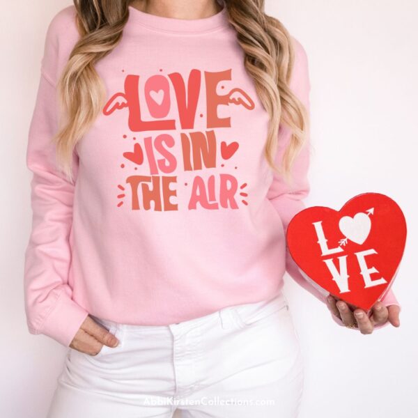A woman, photographed from the neck to the hips. wears a pink sweatshirt while holding a red heart craft. The pink sweatshirt has "love is in the air" printed on it. 