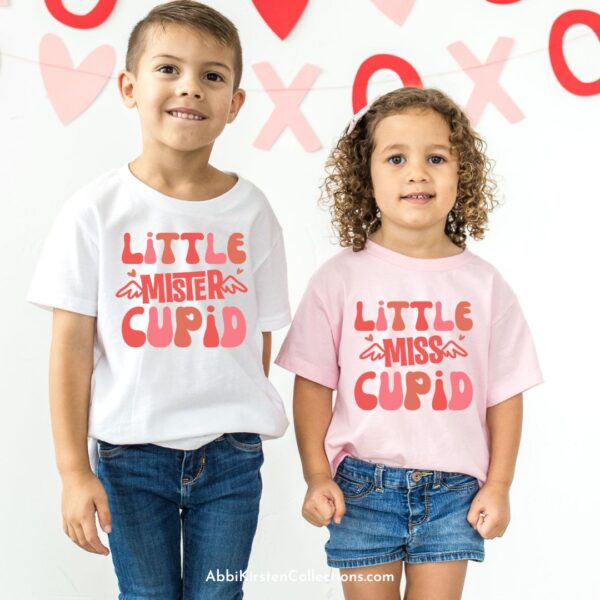 A young boy wearing a white t-shirt that reads "little mister Cupid," and young girl wearing a pink t-shirt that says "little miss Cupid," stand against a white background with Valentine's Day decorations. 