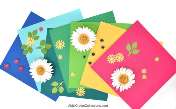 The image shows colorful cardstock with paper flowers on a desk. 