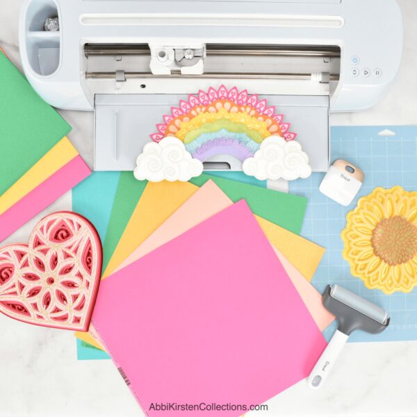 The image shows colorful cardstock next to a Cricut machine with several paper craft projects cut out. 