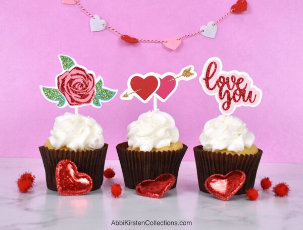 The image shows 3 white cupcakes with handmade cupcake toppers for valentine's day. 