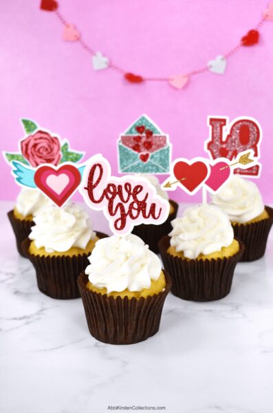 The image shows white cupcakes with handmade Valentine cupcake toppers made with a cutting machine like Cricut. Follow this tutorial and download seven Valentine's Day SVG files for making your DIY cupcake toppers. 
