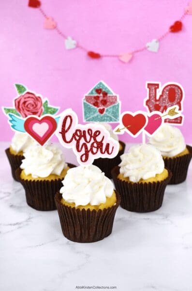 The image shows white cupcakes with handmade cupcake toppers. Create DIY cupcake toppers with your Cricut machine. Follow this tutorial and download seven valentine's day SVG files for making your own cupcake toppers.