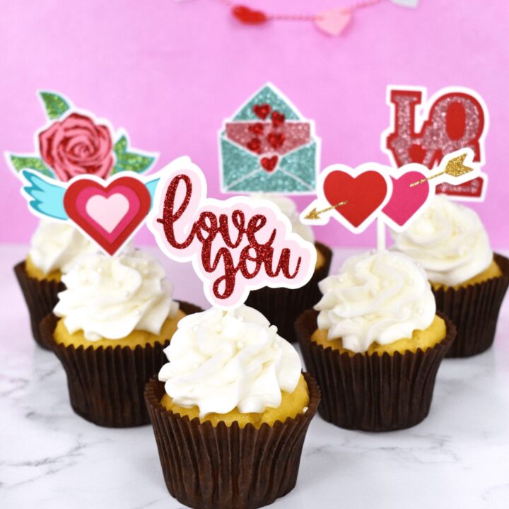 The image shows white cupcakes with handmade cupcake toppers. Create DIY cupcake toppers with your Cricut machine. Follow this tutorial and download seven valentine's day SVG files for making your own cupcake toppers.