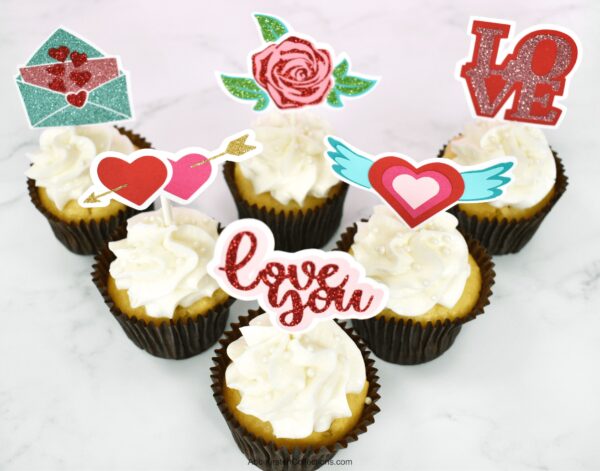 A set of six white cupcakes in a triangle formation each has a different Valentine's Day cupcake topper made with a Cricut machine.  The toppers are two hearts pierced by an arrow, a glittery rose and greenery, an open envelope with hearts, the word “Love,” a heart with wings, and the text “Love you.” 