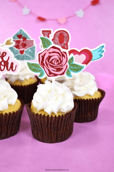 A close-up of DIY cupcake toppers made with Cricut stuck in the top of white-frosted cupcakes with Valentine’s Day decorations. The first cupcake shows a rose design made of cardstock and iron-on vinyl.  