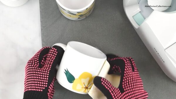 Abbi’s hands, protected by pink heat-safe gloves, peel the backing paper off the freshly pressed sublimation mug blank. Another finished mug with yellow flowers and the Cricut Mug Press sit in the background. 