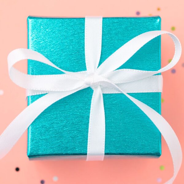 A bird's eye view of a shiny teal-wrapped mystery box has a white ribbon with a simple bow on top. The box rests on an orange table top.