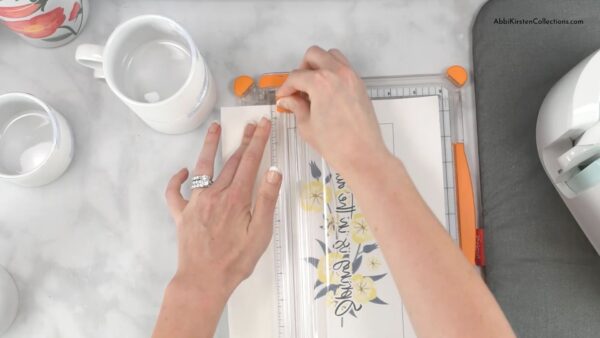 An overhead view of Abbi Kirsten's hands as she uses a straight cutter to trim the excess off the flower design. A heat mat and Cricut machine are visible on the right, and sublimation mugs are on her right. 