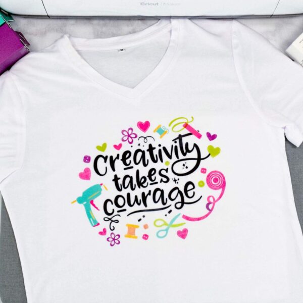 A white t-shirt is laid out on a table neatly. The center has text that says, "Creativity takes courage." The text is surrounded by illustrations of craft supplies like a glue gun and ribbons.