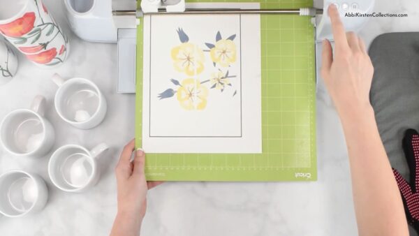 The image shows Abbi Kirsten loading a flower design on sublimation paper into a Cricut machine. The craft table has blank sublimation mugs around the cutting machine. 