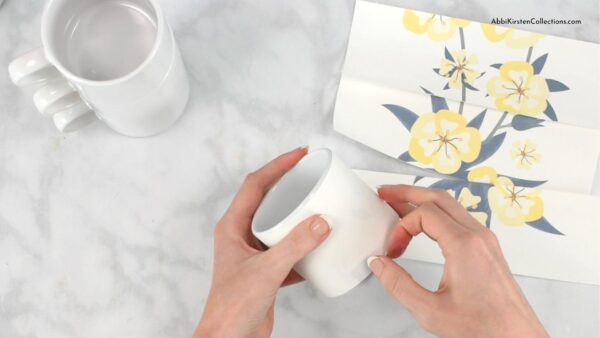 Abbi Kirsten’s hands apply heat-safe tape to the freshly cleaned white sublimation mug. A yellow flower design meant for the mug lies to her right. 