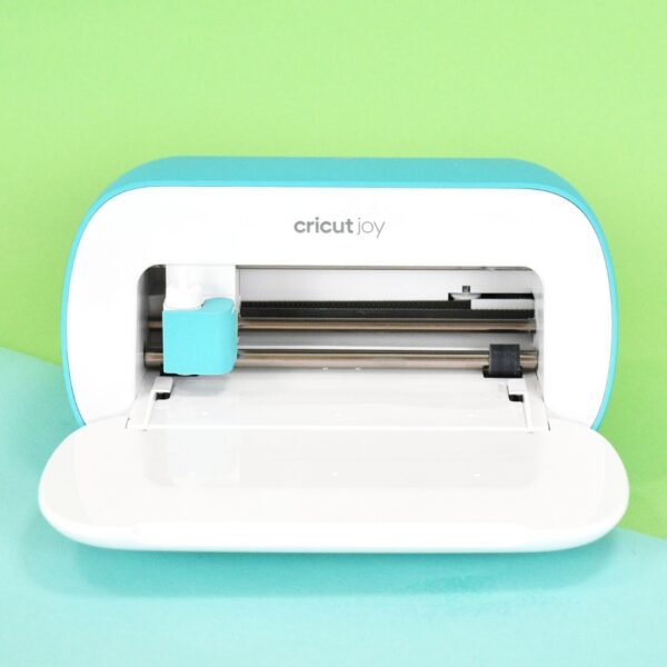 The Cricut Joy machine is open, ready for the mat to be loaded. The cutting machine sits on a light blue surface against a light green background.