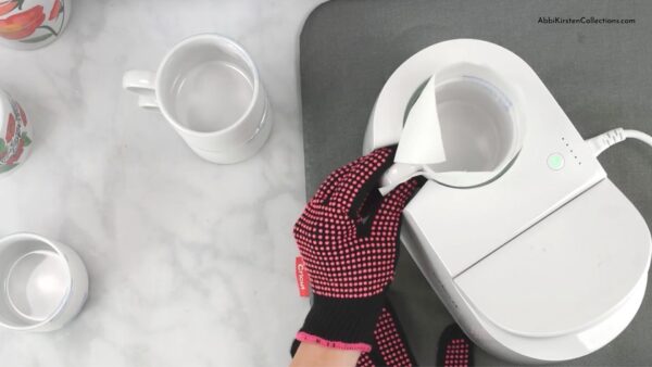 Next to two blank white mugs on the left, Abbi Kirsten has a pink heat-safe glove on her hand as she prepares to use a Cricut Mug Press to add a sublimation design to the current cleaned mug. 