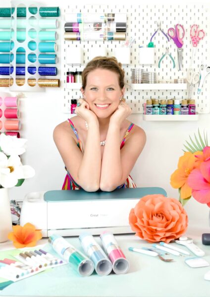 Abbi Kirsten poses in her craftroom with her Cricut cutting machine, surrounded by paper flowers and crafting supplies.