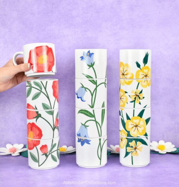 Abbi Kirsten's hand stacks the last mug in a row of three. Each stacked mug has a flower design that, when stacked, creates a larger flower vine. 