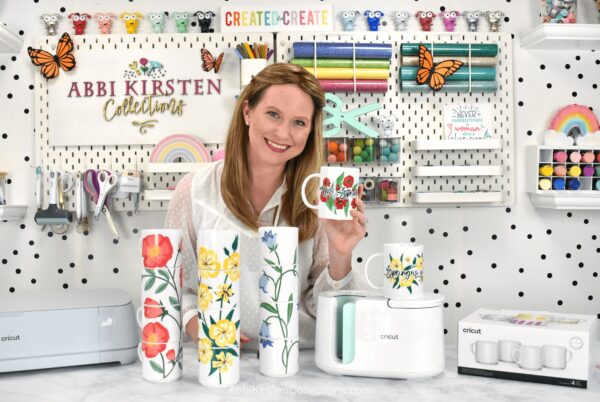 The image shows Abbi Kirsten holding white mugs with spring flower designs while standing in her white, well-supplied craft room. The table in front of her has a Cricut, a Cricut Mug Press, and matching sublimated mugs. 