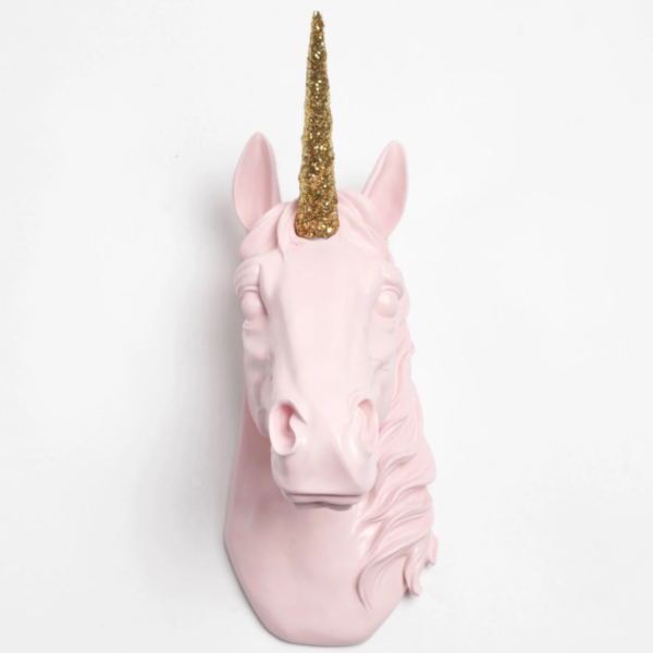 A wall mounted pink unicorn head with a glittery gold horn hangs on a white wall.